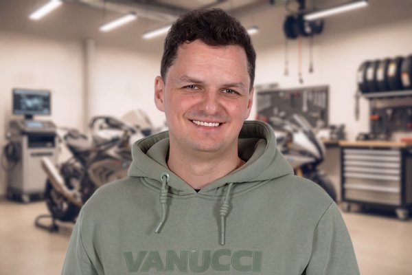 KI generiert: Ein lächelnder Mann in einem grauen T-Shirt mit dem Aufdruck "VANUCCI" hält ein Mikrofon in der Hand. Im Hintergrund sind unscharfe Regale mit verschiedenen Gegenständen zu erkennen.