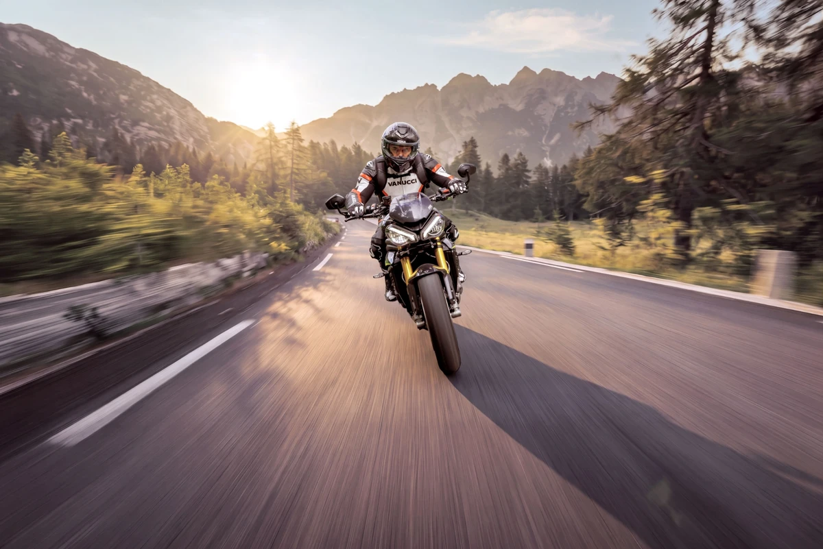 KI generiert: Ein Motorradfahrer fährt auf einer kurvigen Straße durch eine bergige Landschaft bei Sonnenuntergang. Im Hintergrund sind bewaldete Hügel und ein klarer Himmel zu sehen.