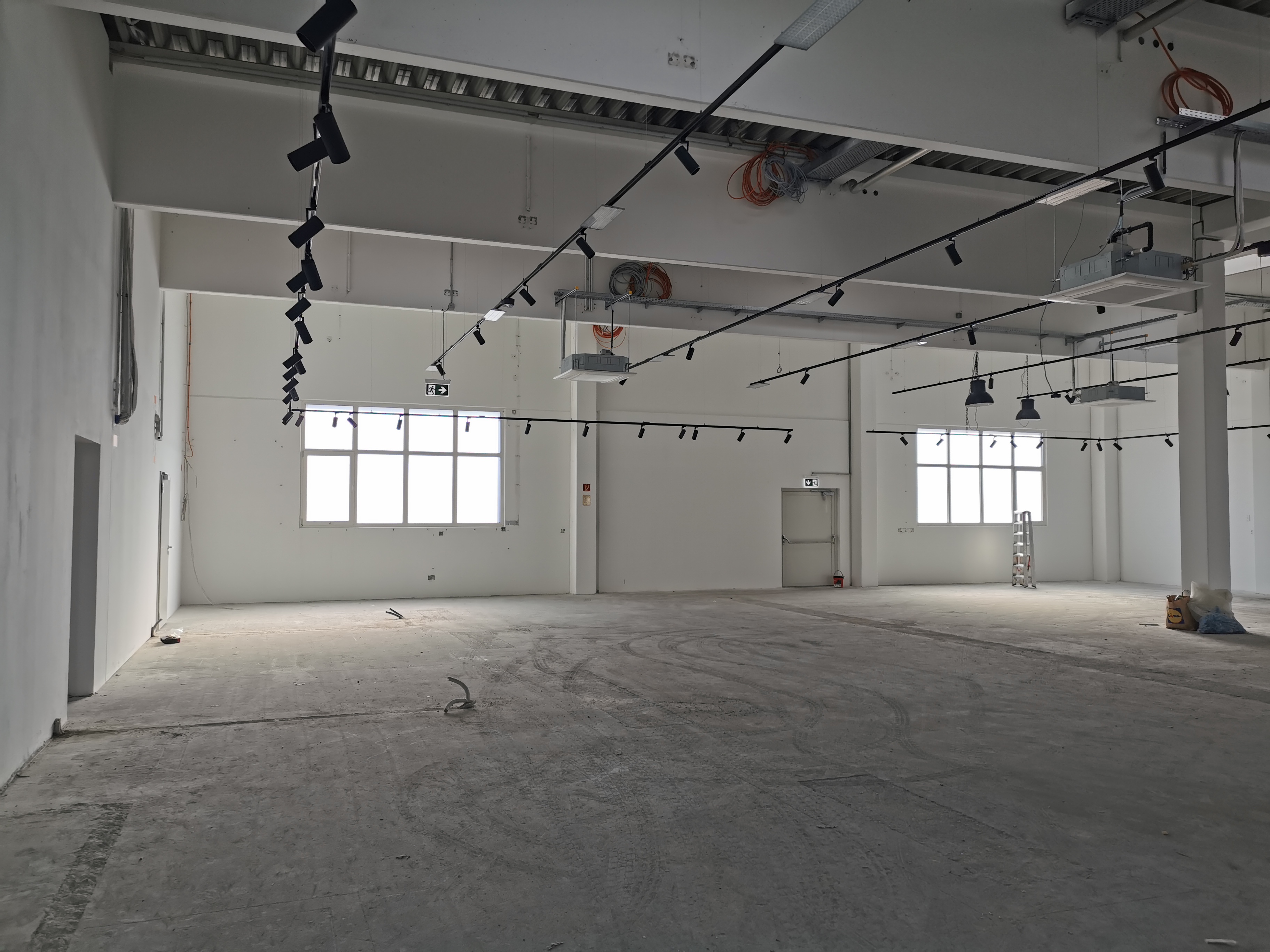 KI generiert: Leerer, unfertiger Raum mit Betonboden, weißen Wänden und großen Fenstern, in dem Elektroinstallationen und Beleuchtungskörper von der Decke hängen.