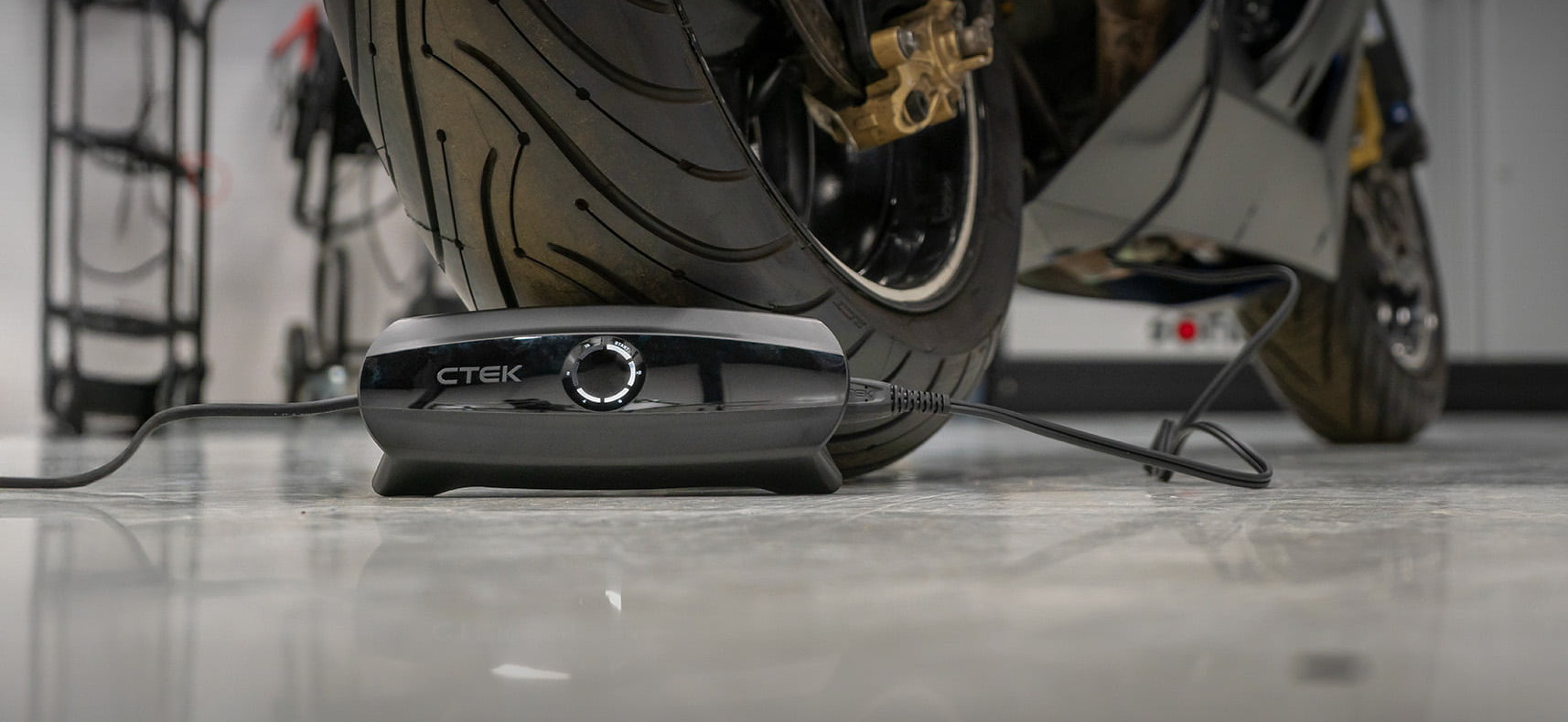 Nahaufnahme eines CTEK‑Batterieladegeräts, das vor dem Hinterrad eines Motorrads auf dem Werkstattboden liegt und angeschlossen ist.