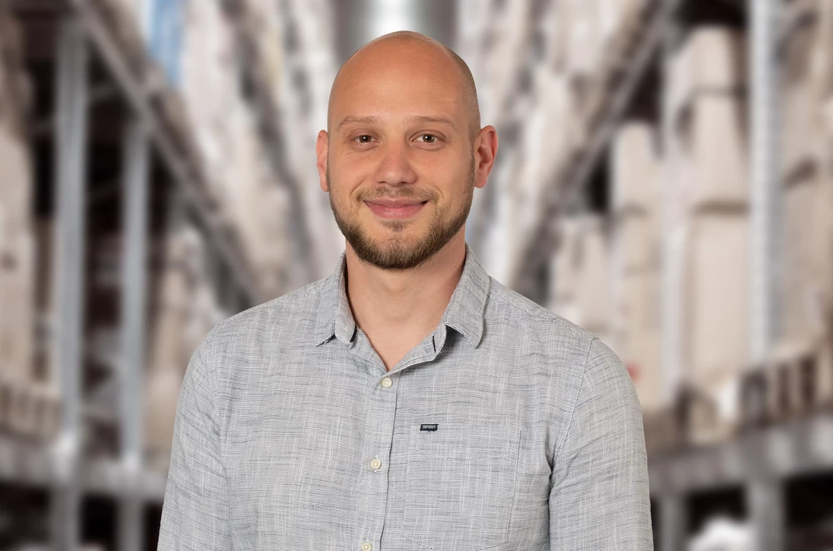 KI generiert: Das Bild zeigt einen lächelnden Mann mit kurzem, dunklem Haar und einem Bart, der ein graues, kariertes Hemd trägt. Im Hintergrund sind unscharf Gebäude und Bäume zu erkennen, was auf eine urbane Umgebung hindeutet.