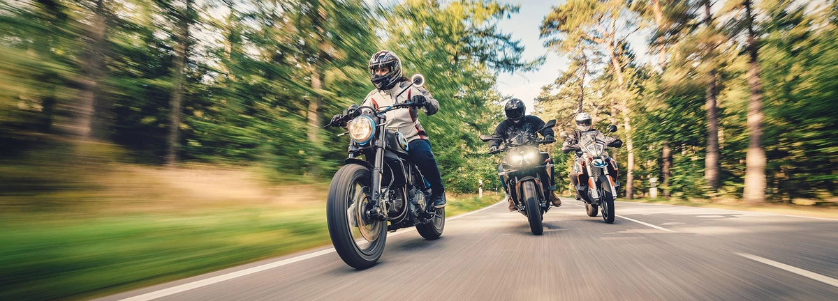 KI generiert: Das Bild zeigt eine Person auf einem Motorrad, die auf einer kurvigen Straße durch eine tropische Landschaft fährt. Im Hintergrund folgen zwei weitere Motorradfahrer auf der gleichen Strecke.