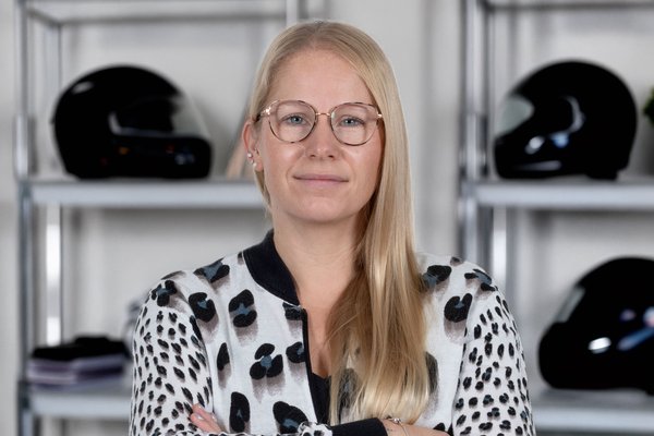 KI generiert: Eine lächelnde Frau mit blonden Haaren und Brille steht vor einem Regal mit Schallplatten. Sie trägt ein gestreiftes Oberteil und eine dunkle Weste.