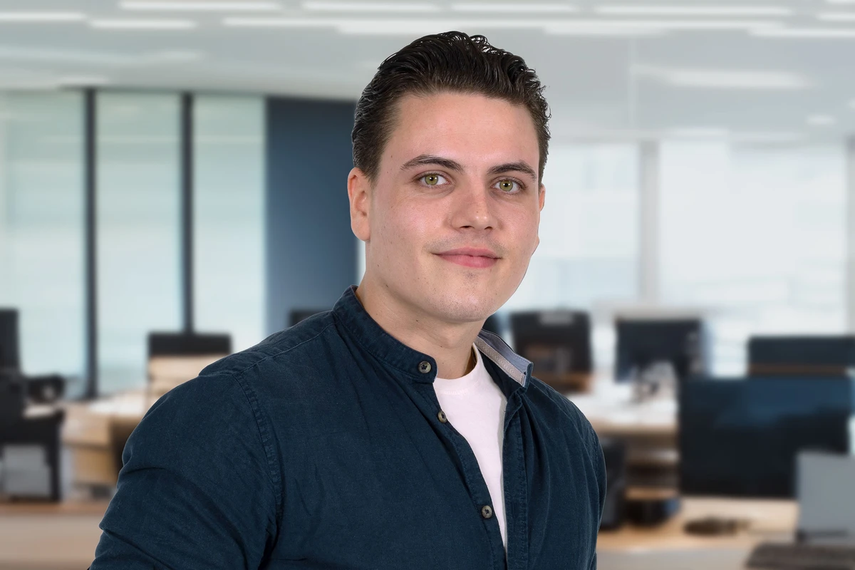 KI generiert: Ein junger Mann mit kurzen dunklen Haaren trägt ein blaues Hemd und einen dunklen Anzug. Er steht vor einem Büro mit Glaswänden und lächelt leicht in die Kamera.