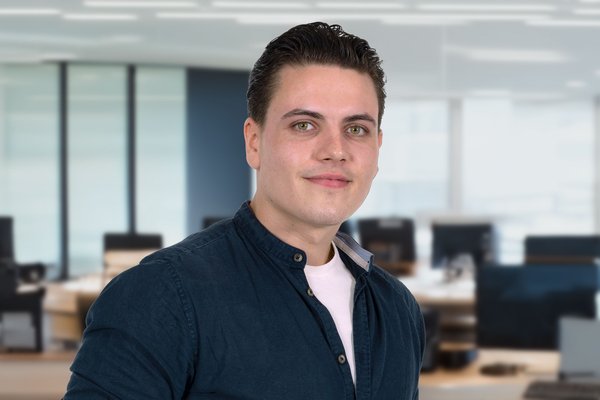 KI generiert: Ein junger Mann mit kurzen dunklen Haaren trägt ein blaues Hemd und einen dunklen Anzug. Er steht vor einem Büro mit Glaswänden und lächelt leicht in die Kamera.