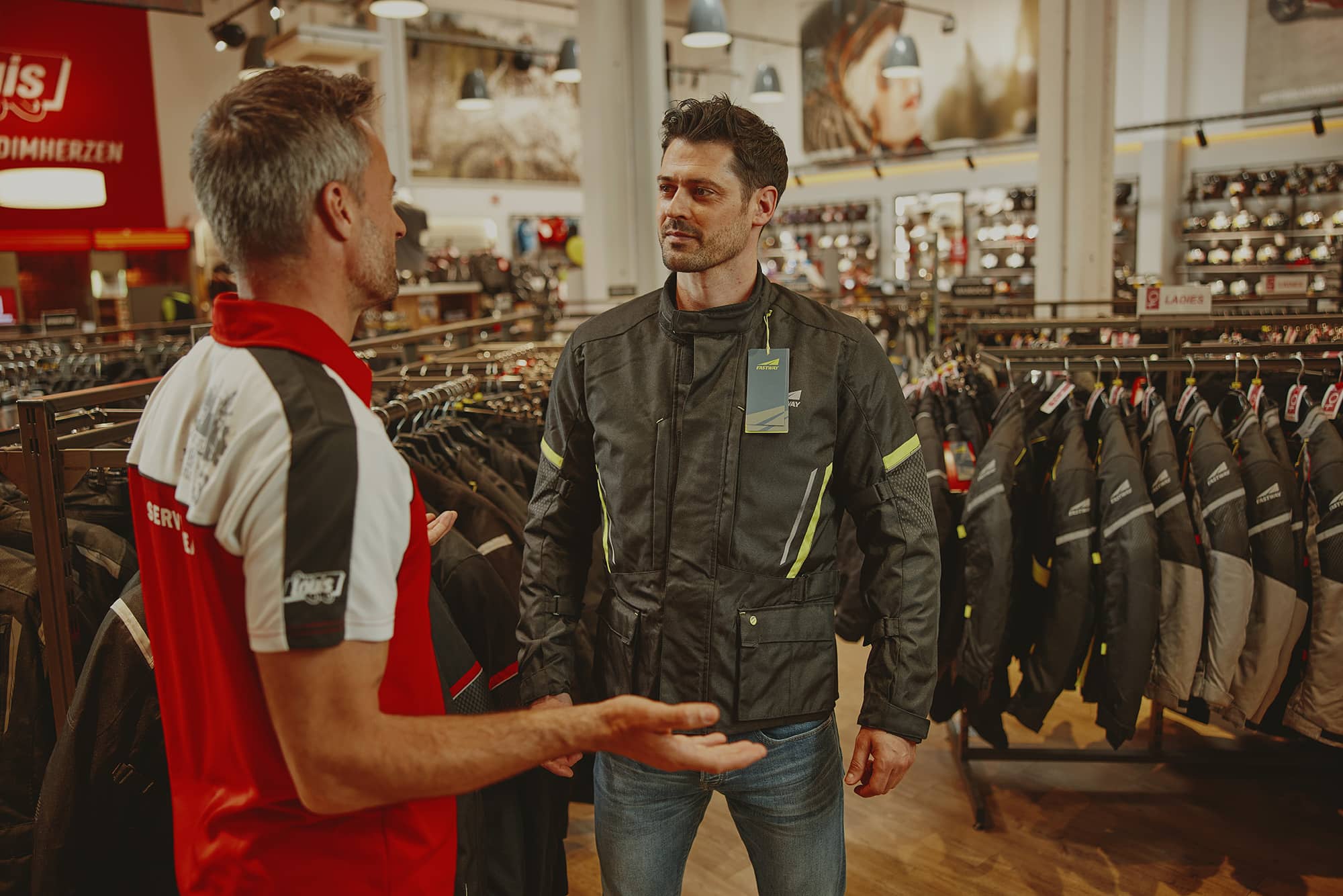 Beratungssituation in einem Motorradgeschäft mit Präsentation von Motorradbekleidung zwischen Verkaufsregalen