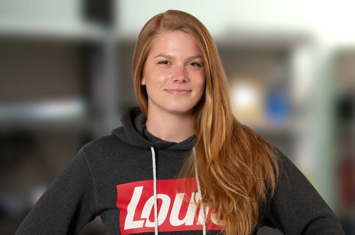 KI generiert: Das Bild zeigt eine lächelnde Frau mit langen roten Haaren, die ein graues Sweatshirt mit dem Aufdruck "Louis" in roten Buchstaben trägt. Im Hintergrund sind unscharfe Elemente zu erkennen, möglicherweise ein Gebäude oder eine Struktur.