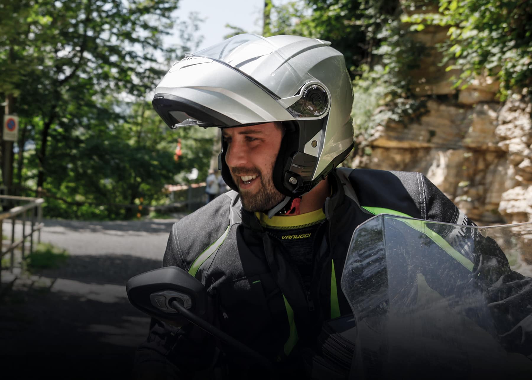 Généré par IA: Un motard portant un casque blanc et une veste verte avec des bandes réfléchissantes, souriant et tenant le guidon de sa moto. Il est assis sur sa moto dans un environnement boisé.