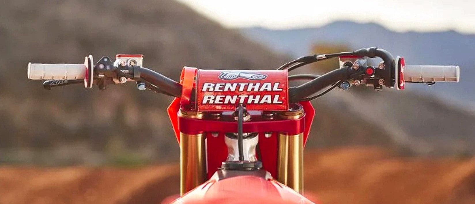 KI generiert: Die Hauptmotive des Bildes sind zwei Motorräder, die auf einer kurvigen Straße durch eine bergige Landschaft fahren. Im Vordergrund ist ein roter Rennlenker mit der Aufschrift "RENTHAL" zu sehen.