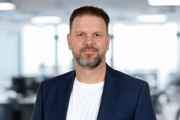 KI generiert: Ein Mann mit dunklem Bart und kurzen Haaren trägt ein weißes Hemd und einen dunklen Anzug. Im Hintergrund sind unscharfe Büroräumlichkeiten mit Glaswänden zu erkennen.