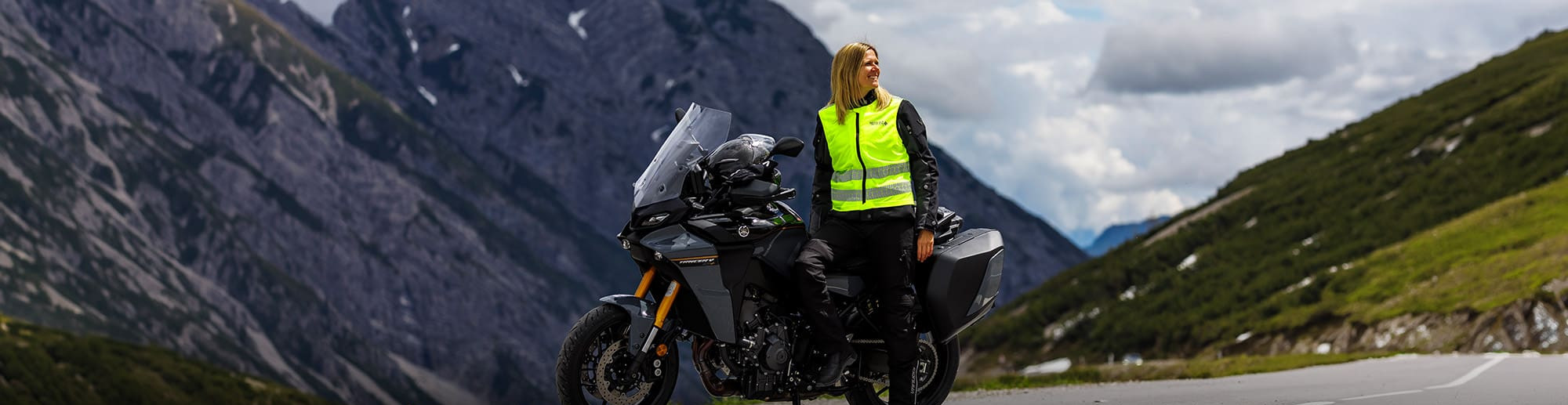 Généré par IA: Un cycliste portant un gilet jaune vif roule sur une route de montagne sinueuse. Il est entouré de paysages alpins spectaculaires avec des sommets enneigés et des prairies verdoyantes.