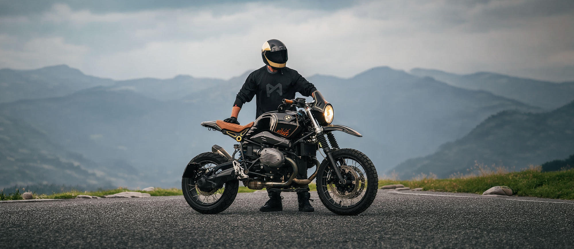 KI generiert: Eine Person auf einem Motorrad fährt auf einer Straße durch eine bergige Landschaft. Im Hintergrund sind schneebedeckte Gipfel und ein bewölkter Himmel zu sehen.