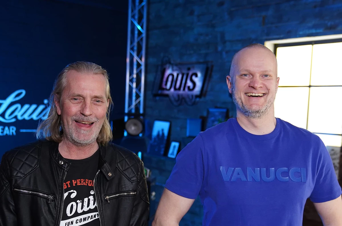 KI generiert: Das Bild zeigt zwei lächelnde Männer vor einem blauen Hintergrund mit dem Louis Vuitton-Logo. Der Mann links trägt ein schwarzes T-Shirt mit der Aufschrift "I ♥ PERFORMANCE", während der Mann rechts ein blaues Polohemd mit dem Namen "VANUCCI" auf der Brust trägt.