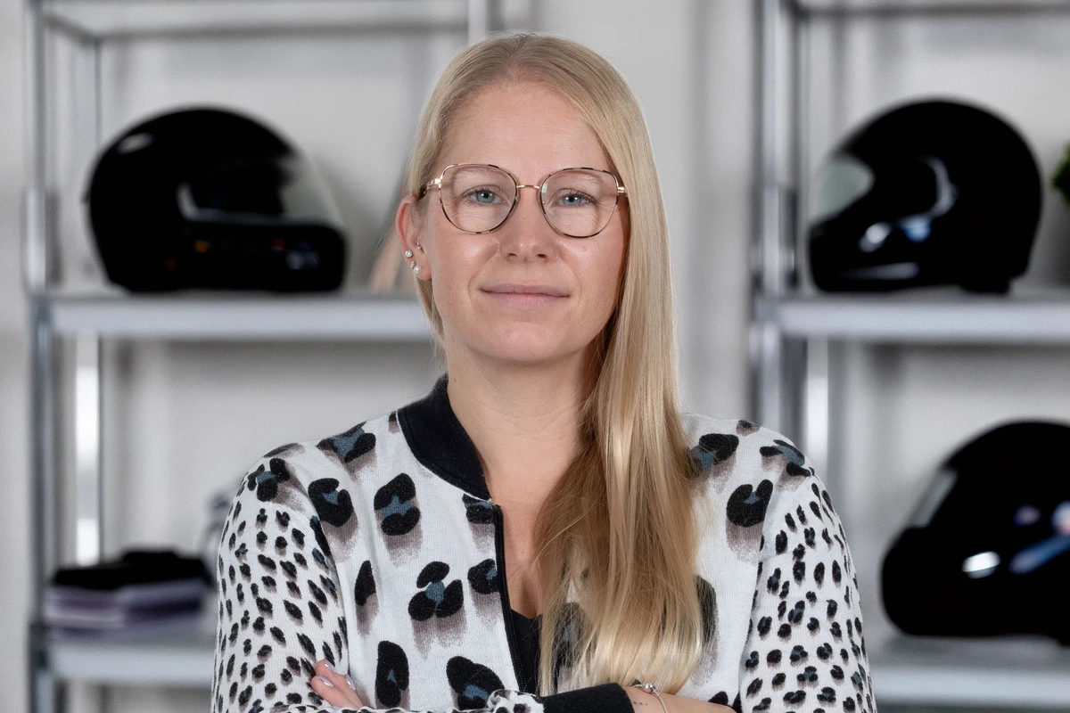 KI generiert: Eine lächelnde Frau mit blonden Haaren und Brille steht vor einem Regal mit Schallplatten. Sie trägt ein gestreiftes Oberteil und eine dunkle Weste.