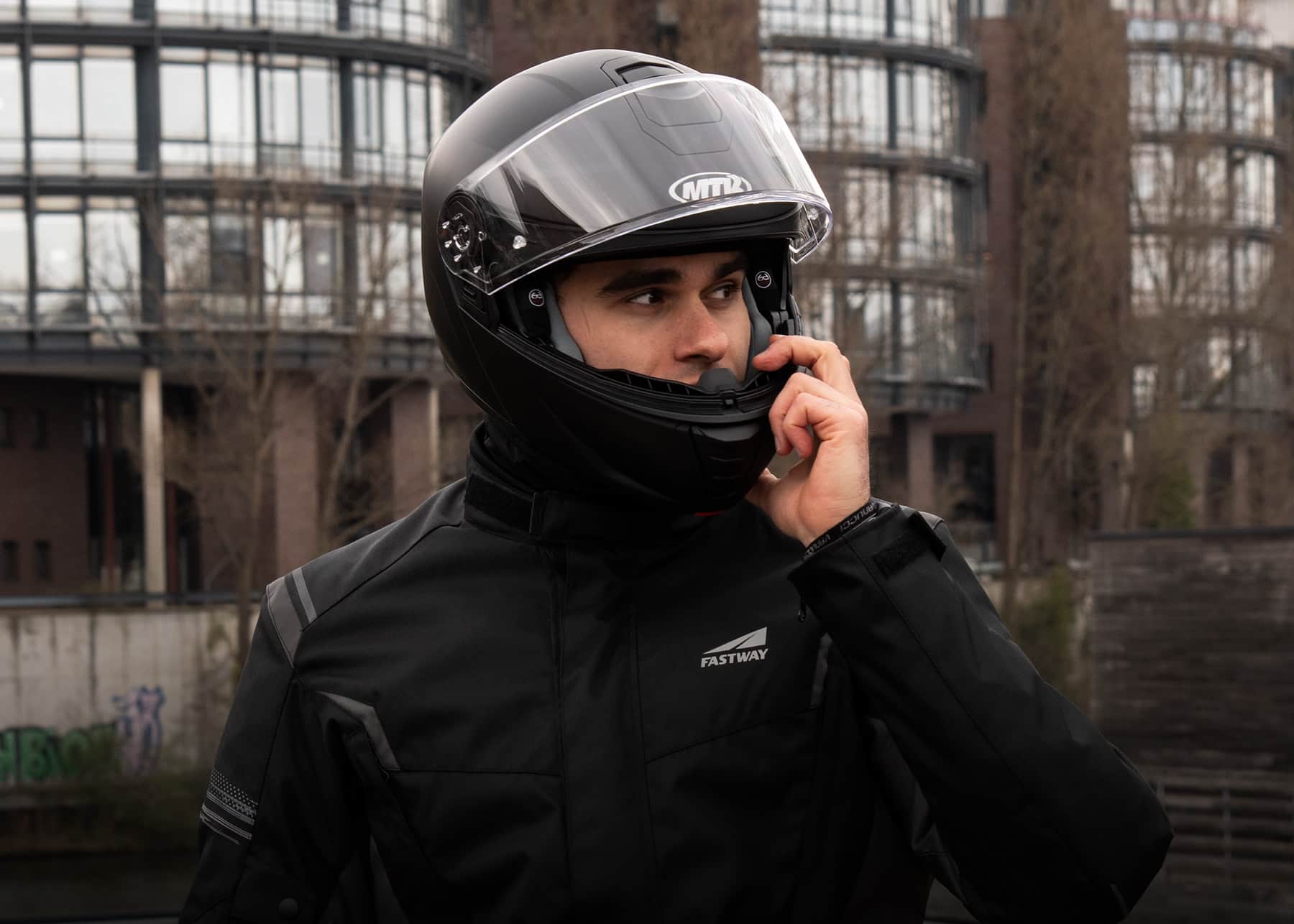Généré par IA: Un motard portant un casque et une veste de protection noire, avec des logos visibles. Il semble communiquer ou saluer de la main gauche.