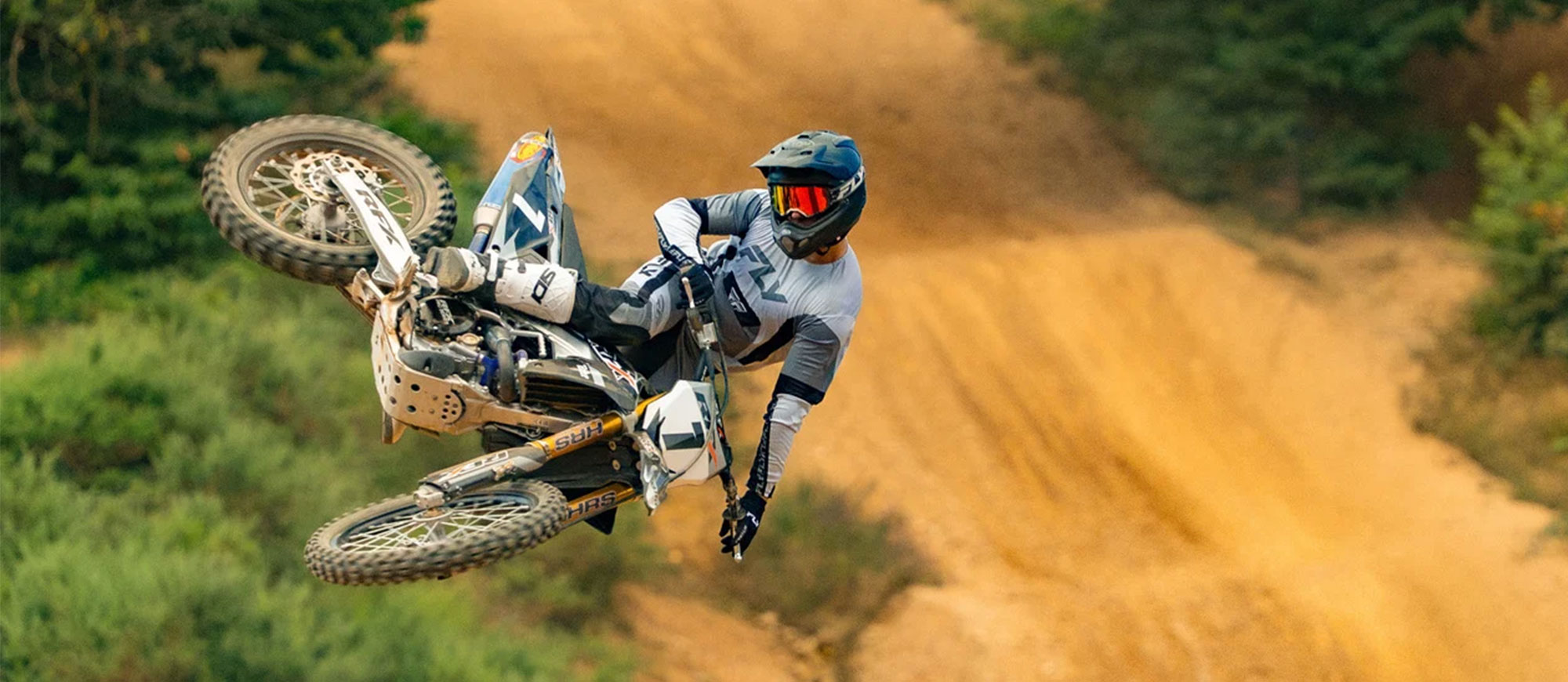KI generiert: Ein Motorradfahrer in voller Schutzausrüstung fährt auf einem Moto-Cross-Motorrad durch eine grasbewachsene Strecke mit einer Staubwolke im Hintergrund. Im Vordergrund ist ein Hund zu sehen, der aufmerksam das Geschehen beobachtet.