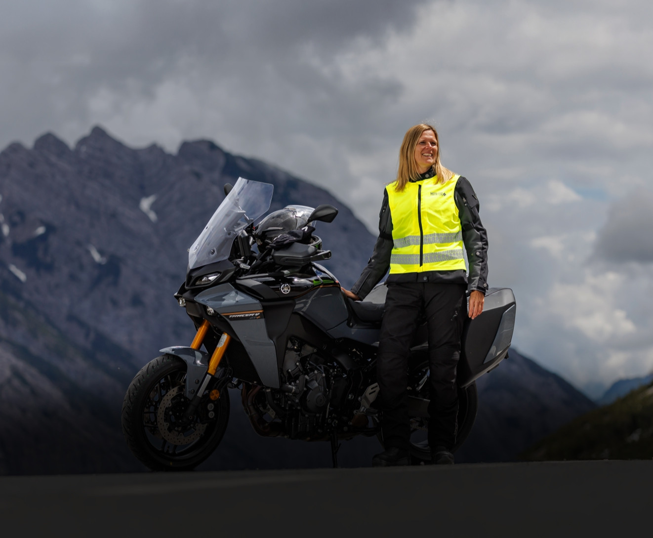 KI generiert: Eine Person in gelber Sicherheitsweste sitzt auf einem Motorrad und blickt in die Ferne. Im Hintergrund sind eine Berglandschaft und ein großer Felsblock zu sehen.